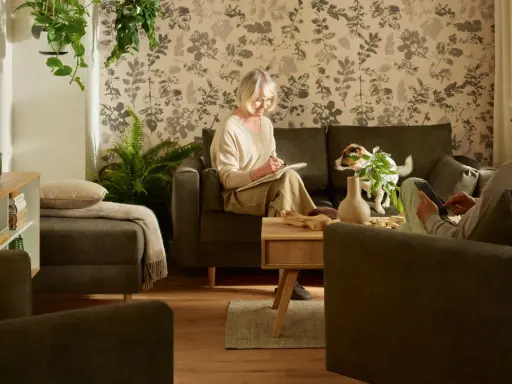 Przytulny salon, gdzie kobieta pisze, a mężczyzna korzysta z tabletu. Idealne inspiracje, jak urządzić pokój dla seniora, z wygodnymi meblami i roślinnością.
